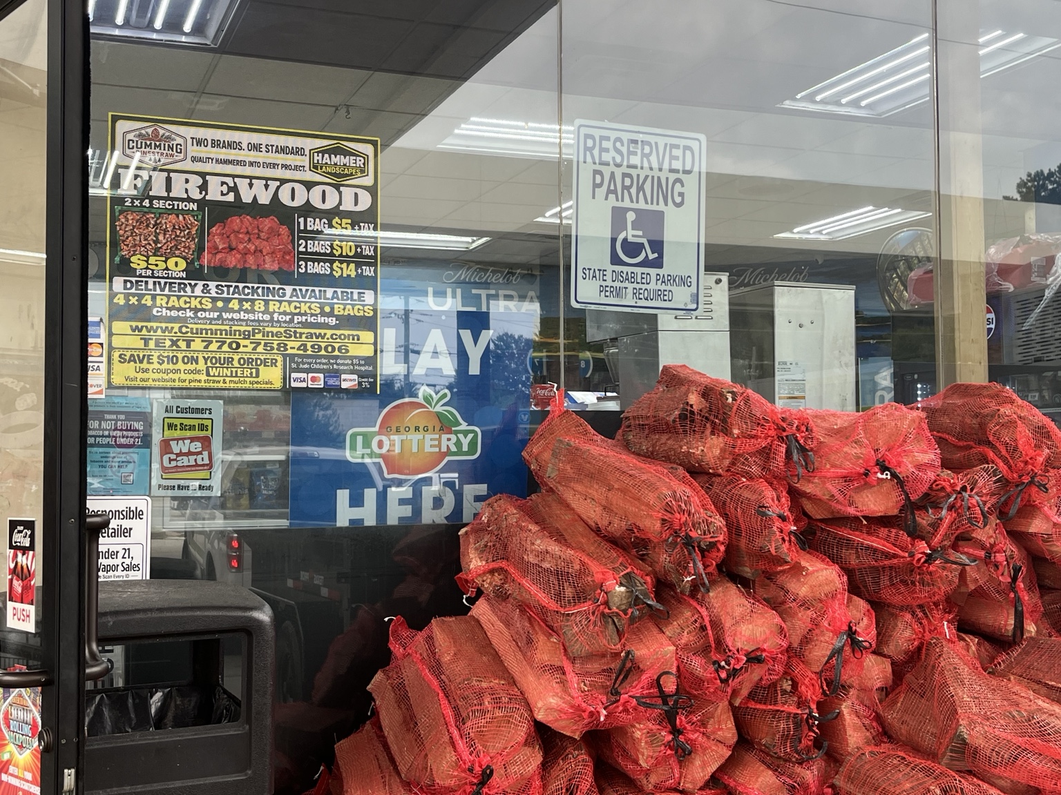 Firewood bags at a partner gas station beneath a window decal displaying Hammer Landscapes and Cumming Pine Straw logos